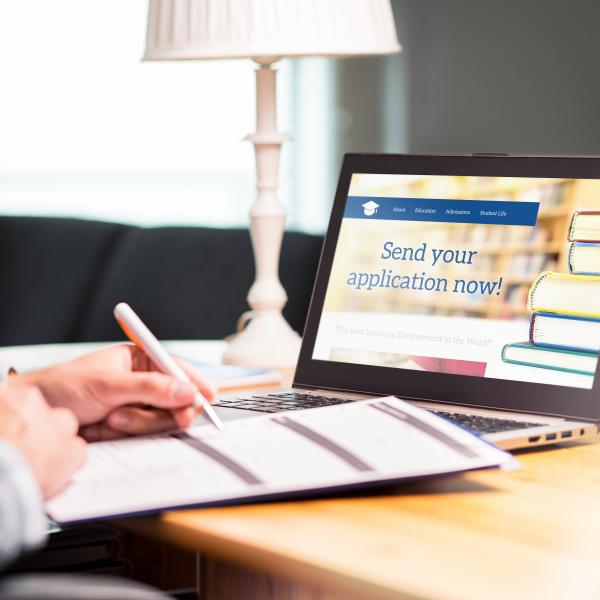 Young man writing college or university application form with pen and paper. Student applying or filling document. Scholarship, grant or admission. School website in laptop.