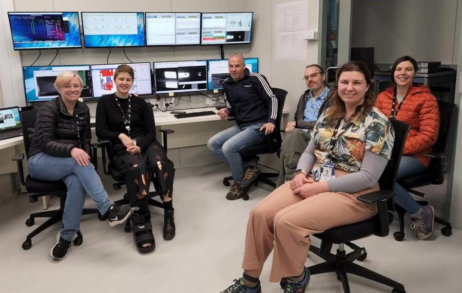 Six scientists sitting in front of a wall of computer monitors. On one of the monitors, rectangles of white light show where the light has entered the beamline.